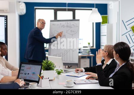 Älterer Geschäftsmann, der einem multiethnischen Team von Kollegen anhand eines Flipchart-Diagramms eine Präsentation gibt. Serious Speaker Chef Executive, Business-Trainer erklärt Entwicklungsstrategie zu motivierten gemischten Rennen Mitarbeiter. Stockfoto