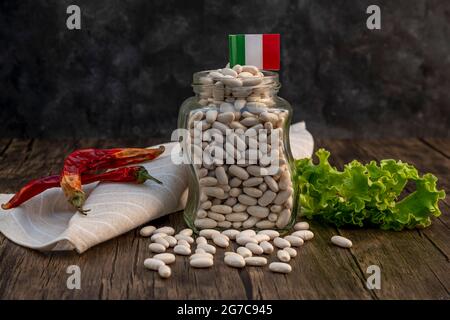 Ein Glas wird mit getrockneten weißen Bohnen gefüllt, auf dem eine italienische Flagge steht, auf einem Tisch mit roten Chilischoten und Salatblättern Stockfoto