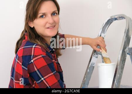 Die Malerin steht auf einer Leiter mit einem Pinsel und einer Dose Farbe, Nahaufnahme. Malen der Wände im neuen Haus Stockfoto