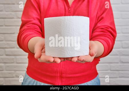 Eine Frau hält eine Rolle Toilettenpapier in Armlänge in Nahaufnahme. Eine Frau steht gegen eine weiße Ziegelwand und hält eine Rolle Toilettenpapier vor h Stockfoto