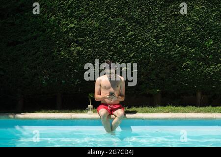 Der junge Mann trägt eine Sonnenbrille und benutzt sein Smartphone, das am Rand des Schwimmbades sitzt. Sommerkonzept. Stockfoto