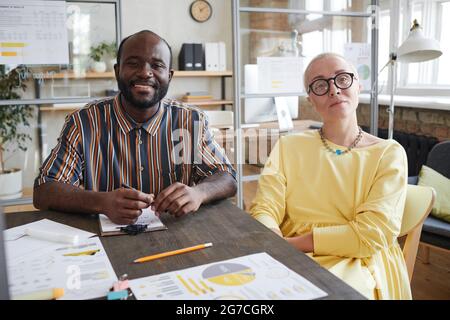 Porträt von zwei Kollegen, die während eines Gesprächs am Tisch mit der Kamera lächeln Stockfoto