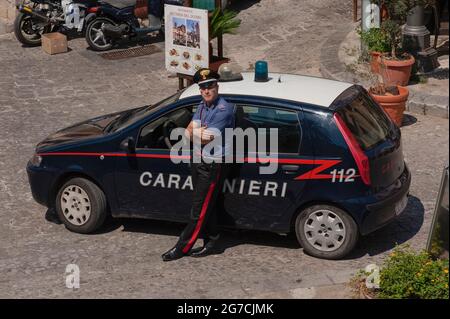 Ich beobachte dich … Ein junger Carabiniere, ein bewaffneter Offizier der italienischen Militärpolizei Arma dei Carabinieri, lehnt sich mit gefalteten Armen gegen sein Fiat-Patrouillenauto, das vor der Via Seminario unter der Vorhofterrasse des Duomo di Cefalu, der historischen Kathedrale in Cefalu, Sizilien, geparkt ist, Und starrt mit stechenden Augen direkt auf die Kamera. Stockfoto