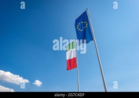 Flaggen der italienischen und der Europäischen Union mit Fahnenmast, die auf einem klaren blauen Himmel mit Wolken und Kopierraum im Wind wehen. Stockfoto