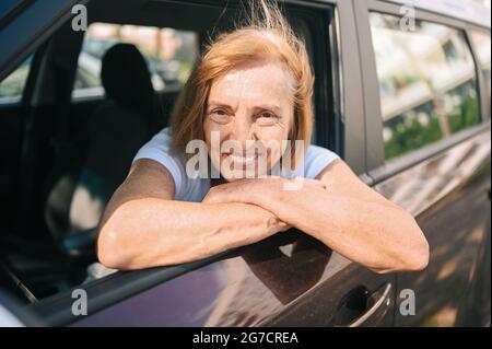 Glückliche ältere Frau, die in einem neuen braunen Auto sitzt, lächelnd auf die Kamera schaut, die die Reise genießt.Fahrkurse und Lebensversicherungskonzept. Aktivitätskonzept für Rentner Stockfoto