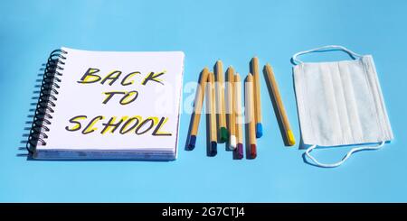 Bleistiftzubehör für Schule und Büro, Notizbuch mit Aufschrift „Back to School“ und medizinischer Maske zur Vorbeugung von Coronaviren, Draufsicht auf blauem Papierhintergrund. Zurück zur Studie während einer Pandemie, hartes Licht. Stockfoto