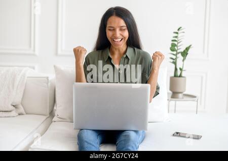 Glück gehabt. Eine begeisterte indische junge Frau feiert Online-Sieg, viel oder geschäftlichen Erfolg, sitzt mit einem Laptop auf der Couch zu Hause und hebt die Fäuste triumphierend Stockfoto