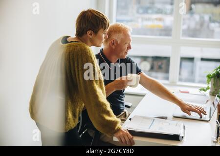 Zwei Geschäftsleute, die im Büro gemeinsam an einem Notebook arbeiten. Geschäftsmann hält Kaffee und zeigt seinem Geschäftspartner etwas auf dem Laptop. Stockfoto