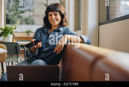 Geschäftsfrau, die auf dem Sofa sitzt und im Büro in die Kamera schaut. Freiberuflicher Mitarbeiter, der im Coworking Space arbeitet. Stockfoto