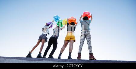 Unkenntlich queere Menschen, die Homosexuell Stolz umarmen. Gruppe von vier Freunden, die im Freien Luftballons mit Regenbogenfarben aufziehen. Junge Mitglieder der LGBTQ+ Stockfoto