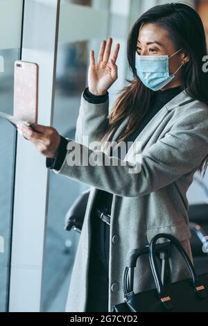 Frau trägt Gesichtsmaske Videoanruf mit ihrem Handy. Weibliche Reisende, die einen Videoanruf in der Wartelounge des Flughafens führen. Stockfoto