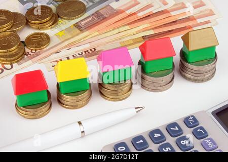 Ein aufsteigender Stapel von Euro-Münzen mit Spielzeughäusern auf der Oberseite, Bargeld, Rechner und Stift. Eine finanzielle Metapher für das Eurohaus. Stockfoto