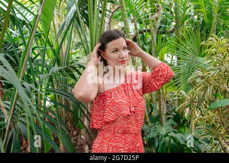 Junge attraktive Millennials-Frau, die in einem sommerlichen Rüschenkleid mit den Händen die Haare hält und von einem üppigen Garten umgeben ist Stockfoto