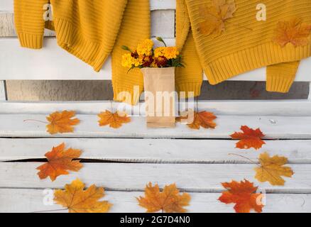 Auf einer weißen Holzbank hängen zwei senffarbene Strickpullover. In der Nähe liegen trockene orange und rote Ahornblätter und ein kleiner Blumenstrauß eingewickelt Stockfoto