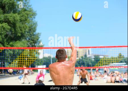 Beachvolleyball. Männlicher Volleyballspieler, der den Ball vor dem Netz serviert, Rückansicht, unscharfe Spieler. Stockfoto