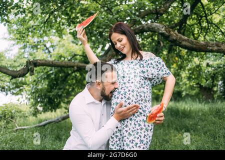 Schöner Mann küsst den schwanger Bauch seiner charmanten Frau an frischer Luft. Hübsche Frau, die aufrichtig lächelt und Wassermelonenstücke in den Händen hält. Stockfoto