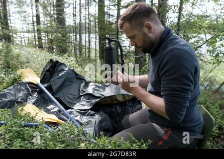 Konzentrierter, ernsthafter Mann hält eine Pumpe in den Händen, um ein Gummiboot zu Pumpen, es gibt Ruder in der Nähe, vor dem Hintergrund eines Sees. Stockfoto