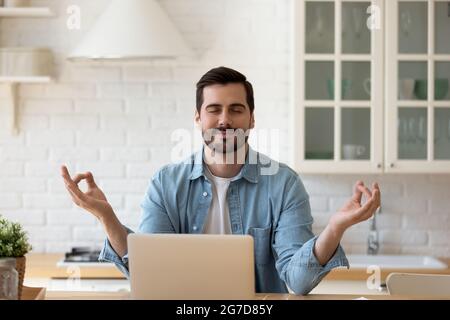 Ruhiger Mann, der in der Küche neben dem Laptop mit geschlossenen Augen meditiert Stockfoto