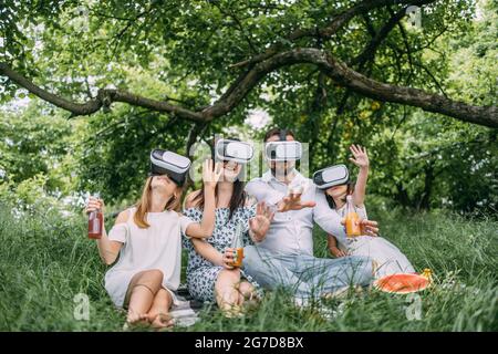 Eltern und ihre beiden Töchter tragen während des Picknicks im Sommergarten ein VR-Headset. Junge Familie, die Spaß mit innovativen Gadgets im Freien. Moderner Lebensstil. Stockfoto