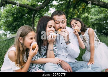 Lächelnde Eltern mit zwei niedlichen Töchtern in Sommerkleidung, die während des Picknicks im grünen Garten hausgemachte Cupcakes essen. Konzept von Menschen, Freizeit und Entspannung. Stockfoto