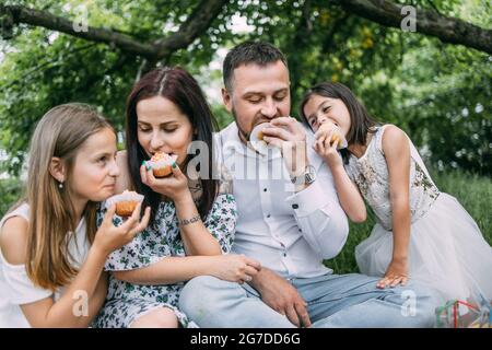 Lächelnde Eltern mit zwei niedlichen Töchtern in Sommerkleidung, die während des Picknicks im grünen Garten hausgemachte Cupcakes essen. Konzept von Menschen, Freizeit und Entspannung. Stockfoto