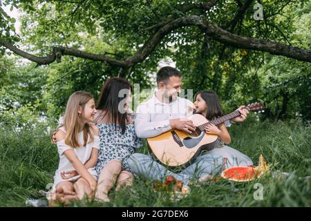 Glückliche Eltern mit zwei Kindern, die auf einer karierten Decke sitzen und während des Picknicks Lieder singen. Vater spielt Gitarre, Mutter umarmt niedliche Töchter. Stockfoto