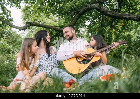 Glückliche Eltern mit zwei Kindern, die auf einer karierten Decke sitzen und während des Picknicks Lieder singen. Vater spielt Gitarre, Mutter umarmt niedliche Töchter. Stockfoto