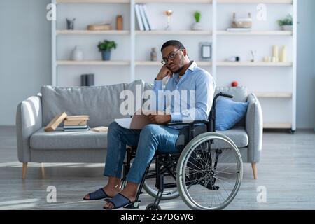 Behinderter schwarzer Mann im Rollstuhl, der Notizbuch liest, von zu Hause aus studiert oder arbeitet, in voller Länge Stockfoto