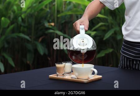 Ein Kellner gießt Kaffee über eine Tasse auf einen mit schwarzem Tuch bedeckten Tisch. Die Atmosphäre im Garten mit verschwommenen grünen Blättern Hintergrund. Stockfoto