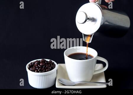 Kaffee aus der französischen Presse-Kaffeemaschine in die Tasse gießen und Kaffeebohnen seitlich, schwarzer Hintergrund, Kopierplatz. Stockfoto