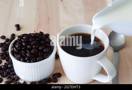 Gießen Sie Milch in die Kaffeetasse, die auf dem Holztisch aufgestellt ist, und legen Sie die Kaffeebohnen zur Seite. Speicherplatz kopieren. Stockfoto