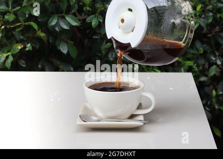 Gießen Sie Kaffee von einer Tasse zu einer Tasse, die auf einem weißen Tisch mit unscharfem grünen Hintergrund steht. Stockfoto