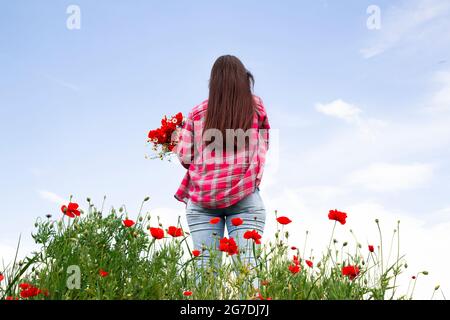 Natur und Feld ein Mädchen mit Blumen in den Händen auf dem Hintergrund wilder Blumen und dem Himmel. Ein Mädchen mit langen Haaren steht mit dem Rücken. Stockfoto