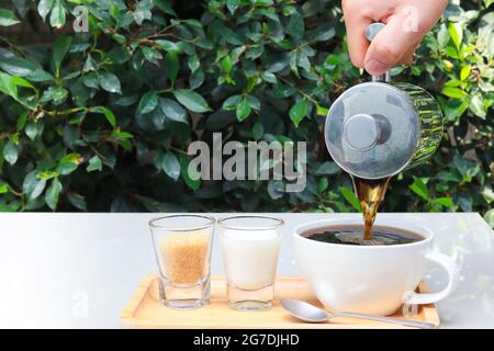 Gießen Sie Kaffee in eine Tasse auf einem Holztablett mit Milch und Zucker daneben. Die Tische befinden sich im Restaurant, im Garten. Speicherplatz kopieren. Stockfoto