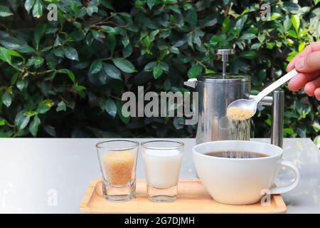 Hinzufügen von Zucker in den Kaffee. Kaffeetasse auf Holztablett und Milch und Zucker daneben. Tisch im Restaurantgarten mit grünen Blättern. Stockfoto