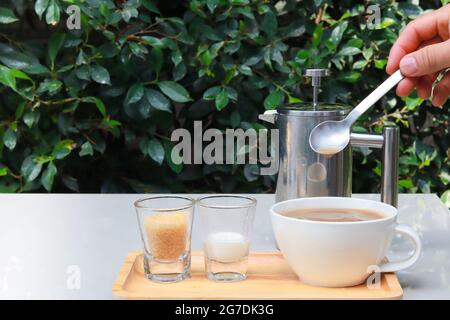 Kaffee, Milch und Zucker auf Holztablett auf Tisch im Garten mit grünen Blättern als Hintergrund und Hand halten Löffel, um Kaffee zu rühren. Speicherplatz kopieren. Stockfoto