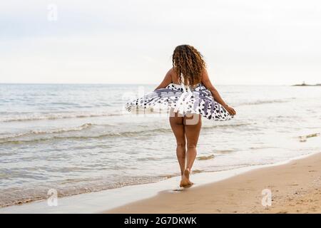 Frau, die bei Sonnenuntergang am Sandstrand entlang geht.Frau, die Sommerferien mit stilvollem Pareo am Meer spielt und tanzt. Stockfoto