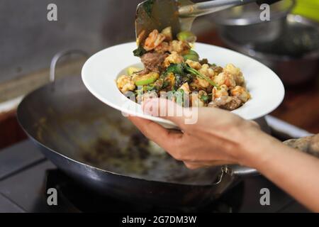 Hände einer asiatischen Frau, die einen Teller und einen Spatel mit Essen vom Wok in den Teller hält. Lokale asiatische Küche Atmosphäre. Stockfoto