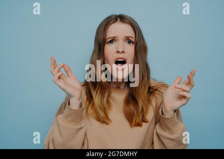 Nettes Teenager-Mädchen, das Unglauben und Schock ausdrückt und gegen die blaue Studiowand steht Stockfoto