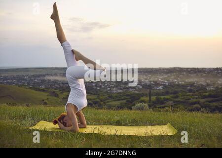 Frau, die bei Sonnenuntergang im grünen Sommerfeld Yoga praktiziert, während sie Kopfstand-Übungen macht Stockfoto