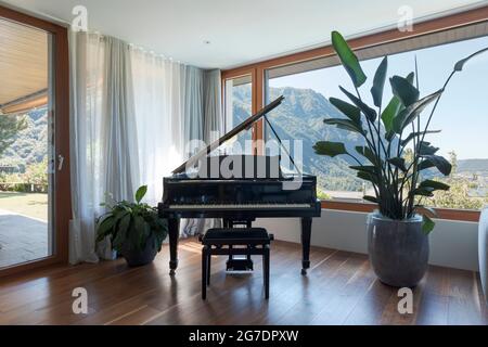 Schwarzes Klavier, Terrasse mit Garten. Großes Fenster mit Blick auf das Tal und den See. Niemand drinnen Stockfoto