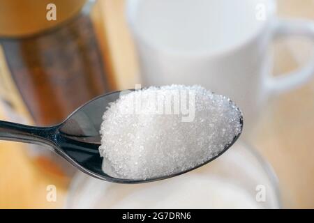 Nahaufnahme eines Löffels Zucker. Löffel mit granuliertem Zucker vor dem Hintergrund der weißen Tasse und des Kaffeejars. Ungesunde Ernährung Konzept. Stockfoto