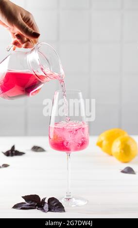 Eine Frauenhand gießt erfrischende rote Basilikumlimonade aus einem Krug in ein Glas. Sommergetränke. Stockfoto