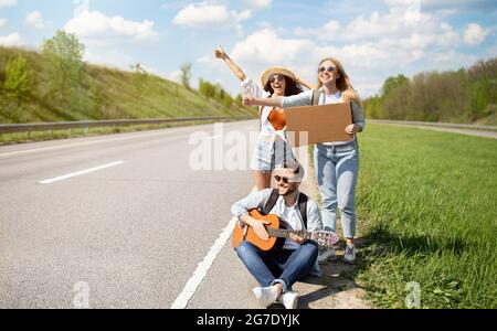 Gruppe junger Freunde, die ihre Fahrt auf der Straße mit leerem Schild, Gitarre spielen, Autostop-Fahrt, freien Platz Stockfoto