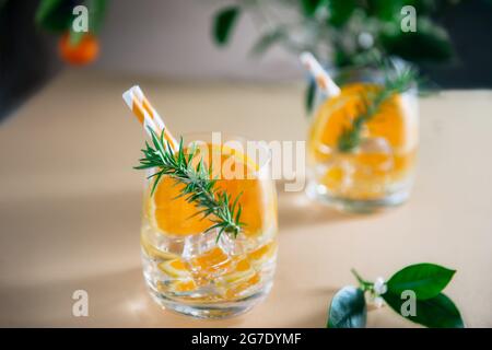 Zwei erfrischende Fruchtcocktails mit Orange, Eis und Rosmarin auf einer neutralen beigen Oberfläche mit einem Hintergrund aus Zitrusbäumen. Getränke an der Sommerbar. Selektiv Stockfoto