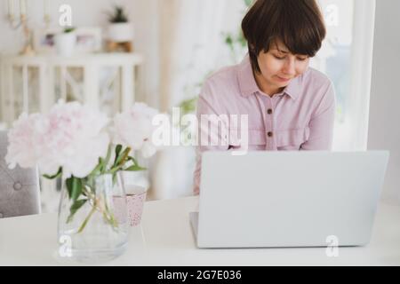 Frau mit gemischter Rasse, die zu Hause mit einem Laptop arbeitet. Gemütlicher Arbeitsplatz mit frischen rosa Pfingstrosen-Blumen und einer Tasse Tee auf dem weißen Tisch mit Licht Stockfoto