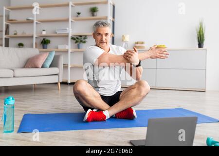 Sport im reifen Alter. Ein glücklicher Mann mittleren Alters, der zu Hause vor dem Laptop Stretching-Übungen macht und auf der Matte sitzt Stockfoto