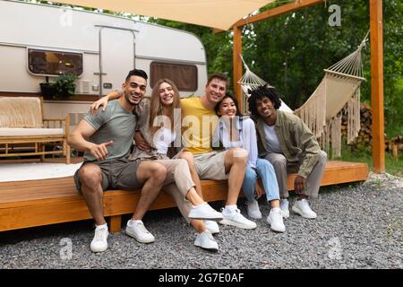 In voller Länge Porträt von multirassischen jungen Freunden, die in der Nähe des Wohnwagens sitzen und die Kamera im Freien anlächeln Stockfoto