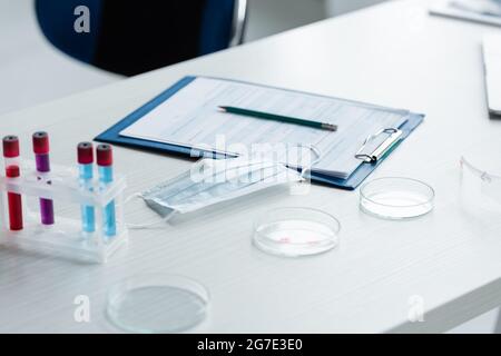 Medizinische Maske in der Nähe von Petrischalen, Reagenzgläsern und Klemmbrett im Labor Stockfoto