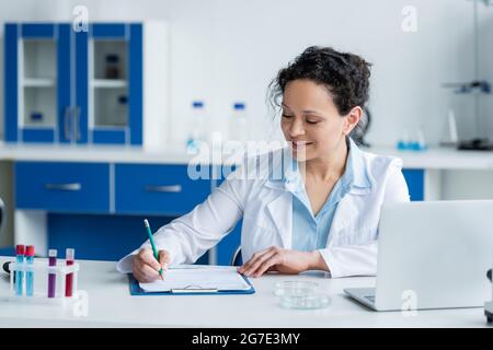 Lächelnder afroamerikanischer Wissenschaftler, der in der Nähe von Laptop und Petrischalen auf die Zwischenablage schrieb Stockfoto